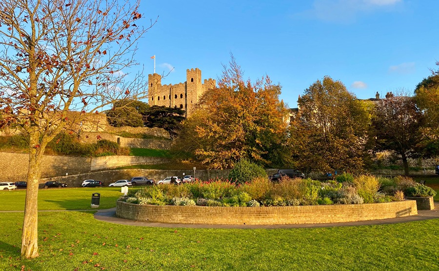 A view of the circular planting bed of the Suffrage Sensory Garden surrounded by grass, with trees as Rochester Castle in the background